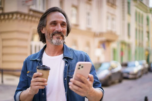 Using navigation mature man stand on street of European city using mobile phone. Travel concept. Handsome middle aged man enjoying his coffee to go traveling in old town wearing casual.