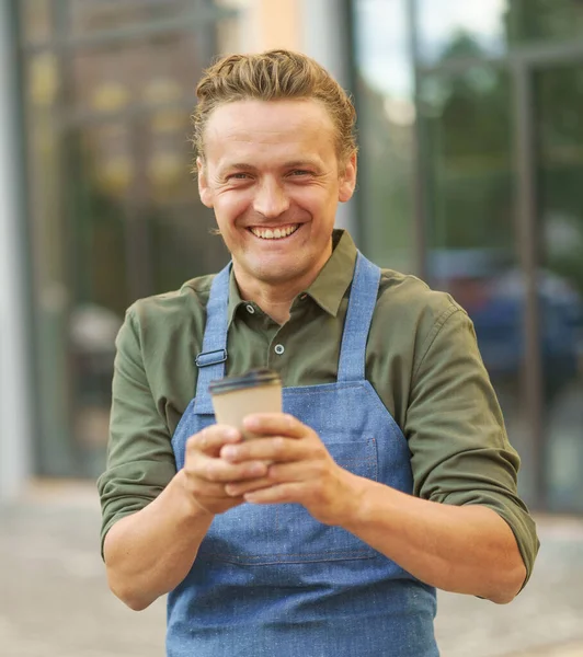 Cheerful handsome barista man with paper cup fresh made coffee gesturing OK sign with other hand and smile standing outdoors with windows shop on background. Freelancer barista in apron.