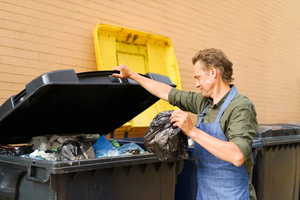 Caucasian handsome man employee open can full of garbage to throw plastic bag with trash wearing apron. Wrong unsorted garbage cans. Wrong way to throw your trash. Sorting garbage save the world.