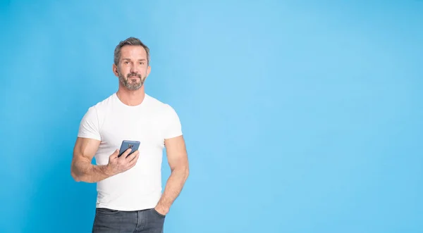 Handsome middle aged grey haired man 40s standing with with smartphone in hand wearing white t-shirt isolated on blue background. Mature fit man, healthy lifestyle concept. Copy space on right.
