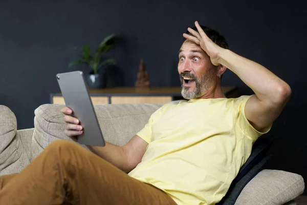 中年のハンサムな男は ビデオ通話やカジュアルな家庭内のソファに座ってショック表情でニュースを読んで通信するためにデジタルタブレットを使用しています フリーランス 自宅から仕事 — ストック写真
