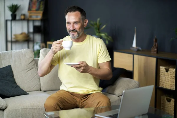 自宅のソファに座ってノートパソコンで作業しながら コーヒーやお茶の中年のビジネスマンをお楽しみください 仕事の距離成熟したItマンはコンピュータで作業しながらコーヒーを飲む — ストック写真