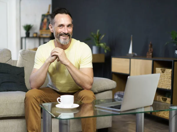 ソファに座っている中年男性の笑顔 ノートパソコンを使って自宅で遠隔作業をしながらコーヒーを飲むソファ 成熟したハンサムなフリーランスItマン仕事上のコンピュータ自宅 — ストック写真