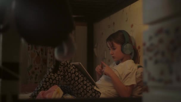 Дитина в навушниках сидить на ліжку і має відеочат через планшет — стокове відео