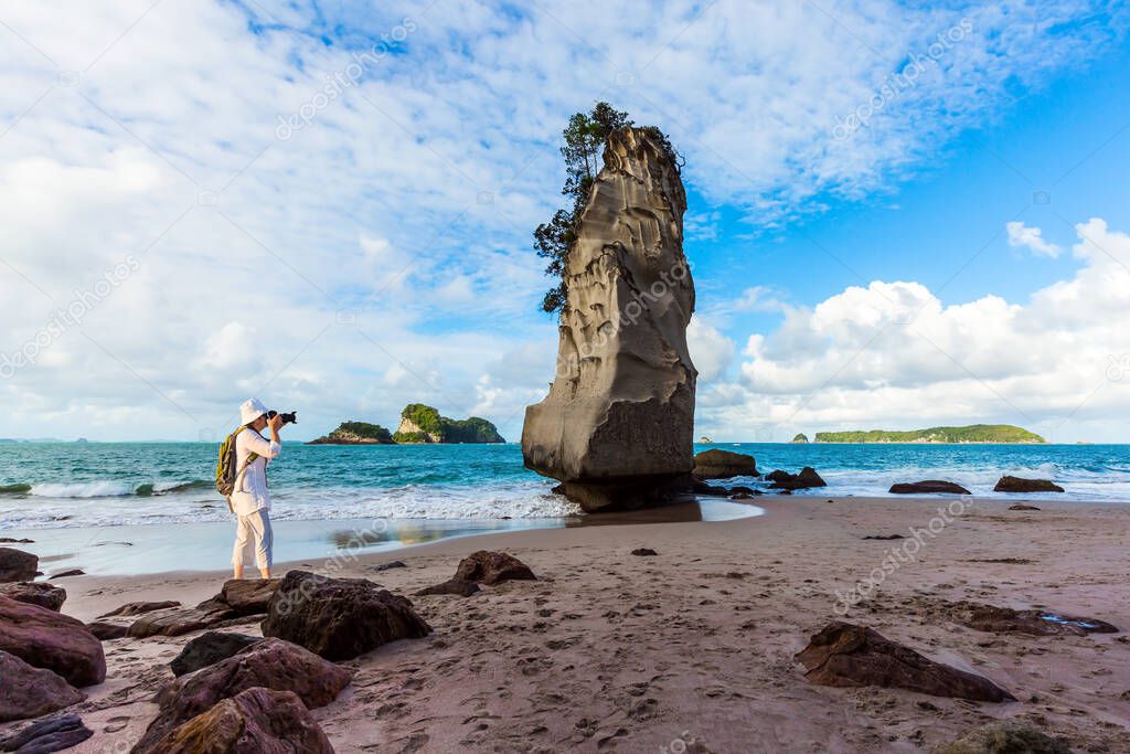 Ocean tide in the Cathedral Cove. Woman tourist taking pictures of beach. Mirror reflections of clouds in wet sand. Travel to New Zealand. The concept of exotic, ecological and photo tourism