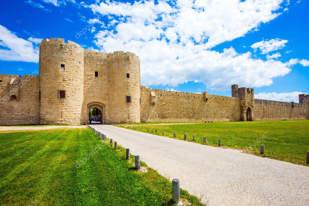  Picturesque powerful gates and fortifications defend the port city of Aigues-Mortes. The concept of historical and photo tourism. Around the walls are green lawns