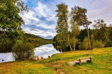 Gölün kenarında rahat bir bank. Resimli buzul gölü Wingdfaller See. Bulutlar ve kıyı ormanı temiz suya çok güzel yansıyor. Almanya, Schwarzwald. Serin bir sonbahar günü