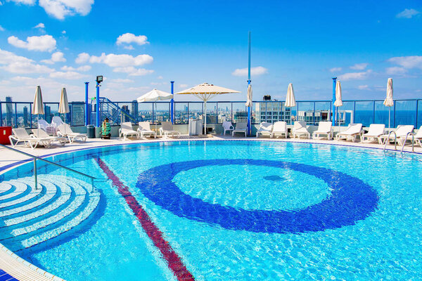 Rooftop pool of a high-rise hotel. The round bowl of the pool is filled with clean, transparent water. There are sun loungers around the pool and waiting for guests