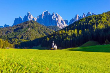 İtalya, Tyrol. Gün batımında yeşil çimlerin arasında çanı olan küçük beyaz bir kilise. Dünyanın en güzel köyü Val di Funes Vadisi 'ndeki Santa Maddalena. Ekolojik ve fotoğraf turizmi kavramı