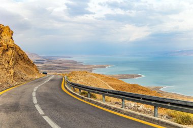 İsrail. Otoyol Ölü Deniz kıyısı boyunca uzanıyor. Denizin karşı kıyısındaki Ürdün loş kış güneşiyle aydınlanır. Bulutlu Ocak günü. Deniz yüzeyi bulutları yansıtır..