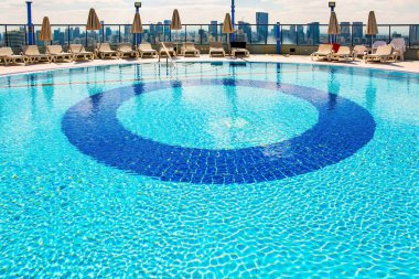 Yüksek katlı bir otelin çatı havuzu. Havuzun etrafında, güneş pansiyonları misafirleri bekliyor. Tel Aviv, İsrail. Havuzun yuvarlak kasesi temiz, şeffaf suyla doludur..