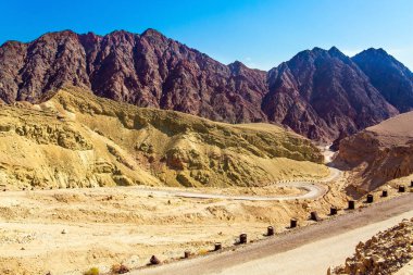Çölün sıcak güzelliği. Kayalar kumtaşlarından, volkanik kayalardan ve volkanik kayalardan oluşur. Eilat Dağları. Toprak yol virajlarda çitle kapatıldı.  