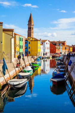 Tekneler kanalların kıyıları boyunca park halindedir. Venedik. Renkli parlak evler ve eğimli bir kule Burano kanallarına yansıyor. Kültür ve fotoğraf turizmi kavramı