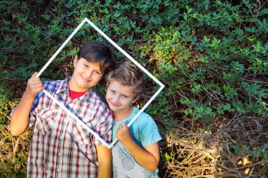 Two adorable boys smiling cheerfully, looking at viewers through a white frame.  Background - green summer buches. Concept - portrait and advertising photo