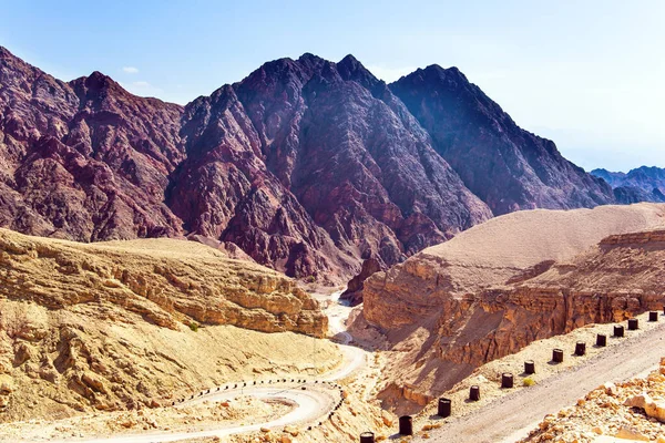 Çölün sıcak güzelliği. Kayalar kumtaşlarından, volkanik kayalardan ve volkanik kayalardan oluşur. Eilat Dağları. Toprak yol virajlarda çitle kapatıldı.  