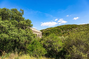 The aqueduct Pont du Gard connects hills covered with dense deciduous forest. The Pont du Gard is the tallest Roman aqueduct. The Gardon River in a bright sunny day. Interesting trip to France. 
