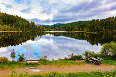 Göl kenarındaki rahat piknik masası ve banklar. Buzul Gölü Wingdfaller See. Bulutlar ve kıyı ormanı berrak sulara yansıyor. Almanya, Schwarzwald. Serin bir sonbahar günü