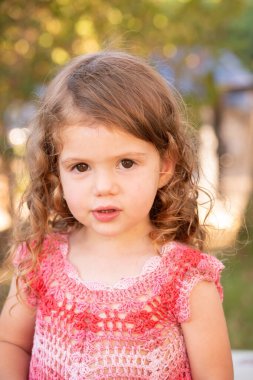 Portrait. Charming little girl with curly hair plays on a green grassy lawn. The girl laughs merrily and claps her hands. Warm spring day. 
