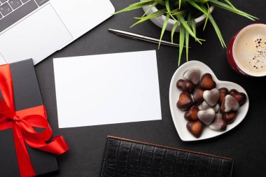 Hediye kutusu ve çikolatalı kalp şekerlemeleri ofis masasında dizüstü bilgisayarın üzerinde. Üst görünüm dairesi boş tebrik kartınızla birlikte yatıyordu. Sevgililer Günü şablonu
