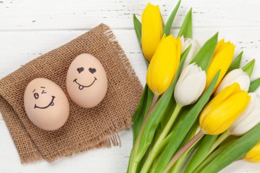 Paskalya tebrik kartı, üzerinde komik yüzler ve ahşap arka planda lale çiçekleri olan paskalya yumurtaları. Üst görünüm düzlüğü