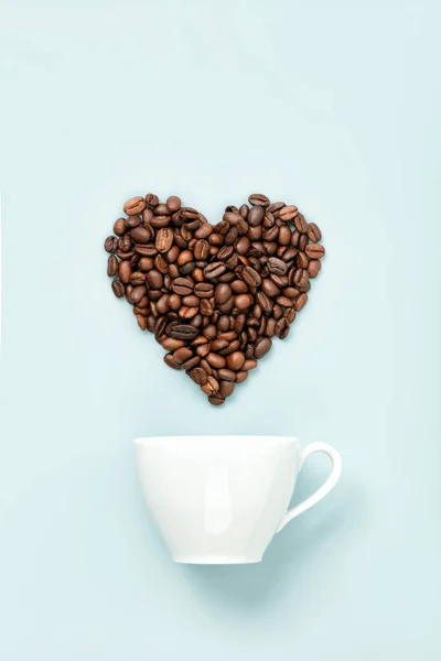 ถ้วยกาแฟสีขาวและถั่วกาแฟในรูปร่างของหัวใจบนพื้นหลังสีฟ้า, แบนวาง — ภาพถ่ายสต็อก