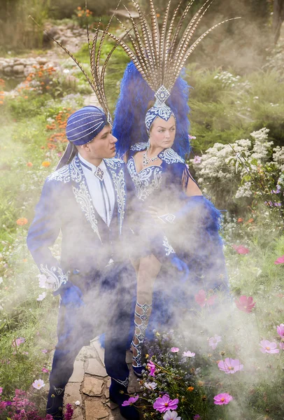 Couple in expensive dark blue costume of illusionist pose in fairy tale flowers park. — Stock Photo, Image
