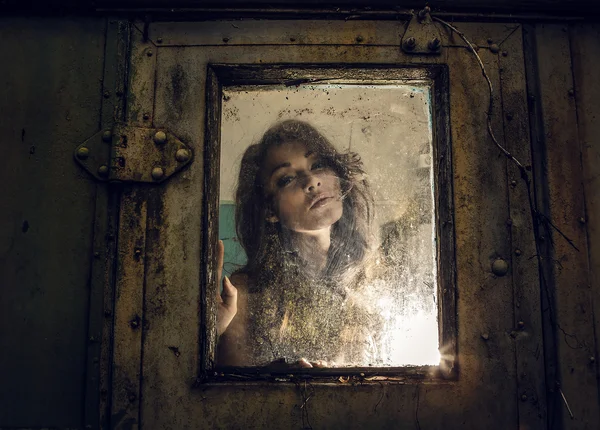 Kunstporträt einer schönen jungen gruseligen Frau, blickt durch ein im Grunge-Stil gestaltetes regnerisches Fenster. — Stockfoto