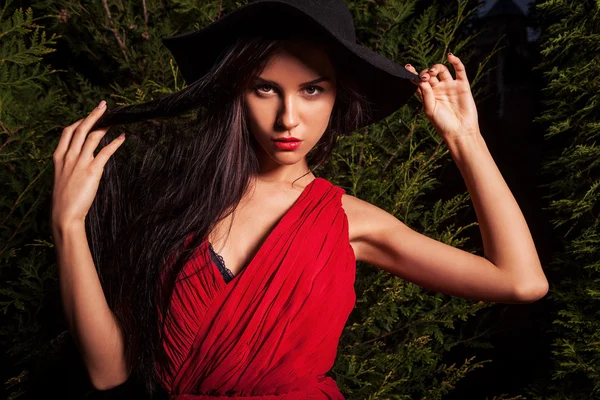 Bruneta Krása ženy v červených šatech & klobouk představují v noci park. — Stock fotografie