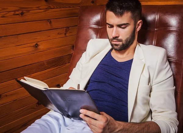 Joven guapo en traje blanco relajándose en un sofá de lujo con diario . — Foto de Stock