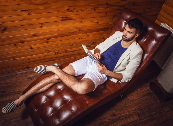 Joven guapo en traje blanco relajándose en un sofá de lujo con diario . — Foto de Stock