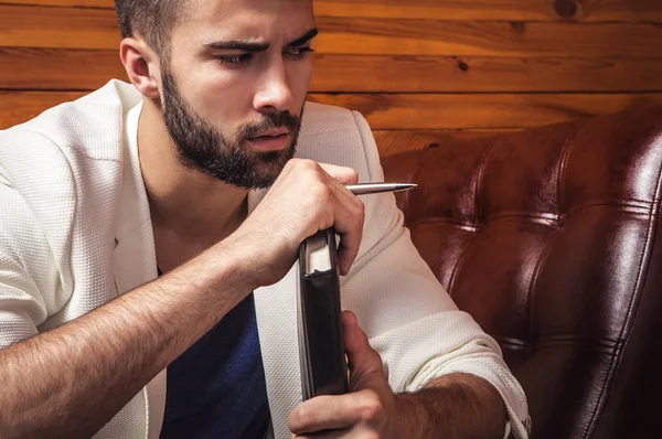 Joven guapo en traje blanco relajándose en un sofá de lujo con diario . — Foto de Stock