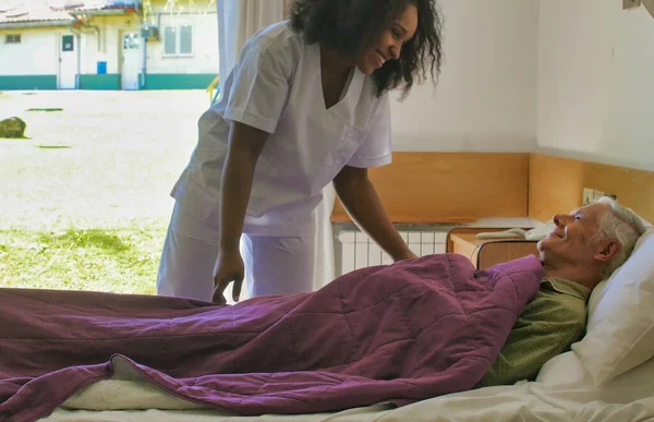 Νεαρή Αφρικανή Γιατρός Που Βοηθά Ηλικιωμένο Συνταξιούχο Στο Κρεβάτι Του — Φωτογραφία Αρχείου