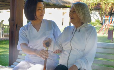 Asyalı doktor, yaşlı ve emekli bir kadınla konuşuyor. İkisi de bahçe bankında oturuyor. Mutluluk ve emeklilik kavramı.