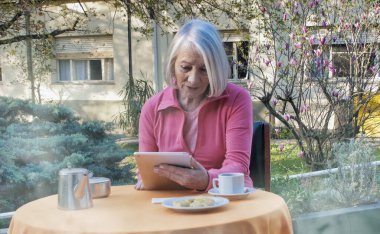 Kafkasyalı emekli kadın kahvaltı için bahçede tablet kullanıyor, video görüşmesi yapıyor. Emeklilik, teknoloji ve rahatlama kavramı.