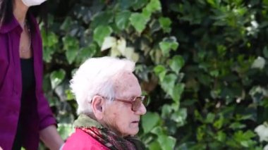 Engelli annesine parkta tekerlekli sandalyede yardım eden genç bir kadının yan görüntüsü.