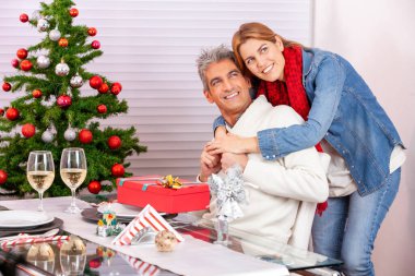 Mutlu gülümseyen genç çift Noel ağacı önünde kucaklaşıyor.