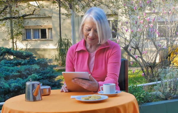 白种人的退休妇女使用科技产品在花园里放松地吃早餐 并有一个视频 技术和放松概念 — 图库照片