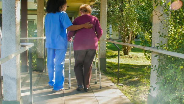 病院の庭にウォーカーの棒を持つ高齢者の女性と話すアフリカの女性医師 幸福とリハビリテーションと退職の概念 — ストック写真