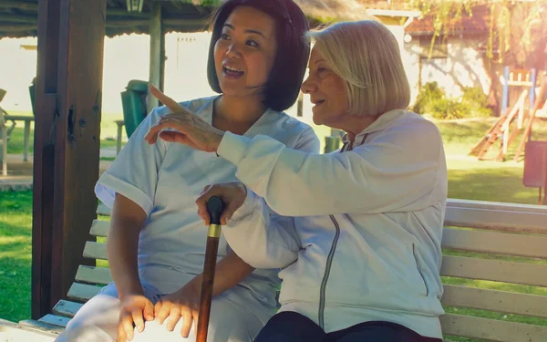 亚洲医生用手杖与年老的退休妇女交谈 她们都坐在花园的长椅上 幸福与退休概念 — 图库照片