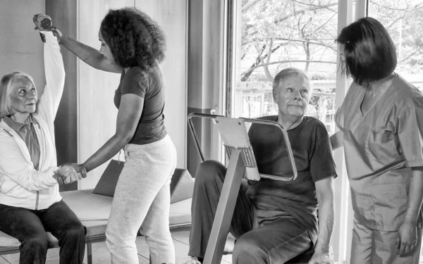 Rehabilitation concept. Elderly retired couple training in a gym supervised by expert african and asian female trainers.