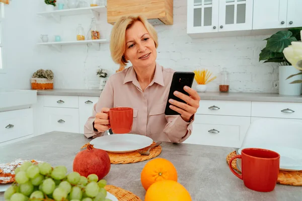 Frau frühstückt zu Hause mit Obst, Kuchen und Kaffee und liest Nachrichten vom Smartphone — Stockfoto