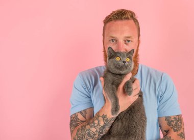 Sakallı ve dövmeli bir adam gri bir kediyi okşar.