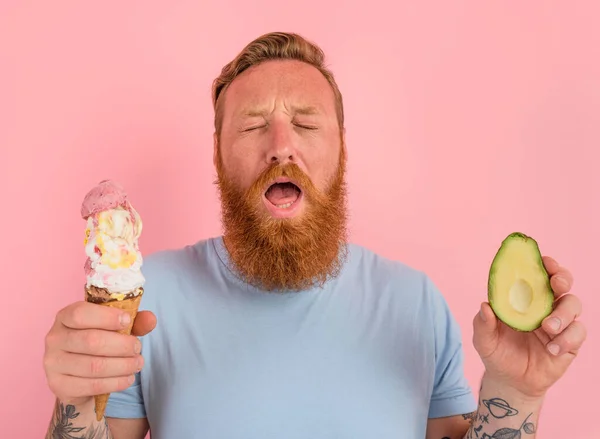L'uomo premuroso con la barba e i tatuaggi è indeciso se mangiare un gelato o un avocado. — Foto Stock