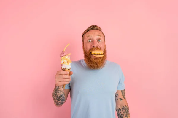 Erstaunter Mann mit Bart und Tätowierungen ist unentschlossen, ob er ein Eis oder ein Sandwich mit Hamburger essen soll — Stockfoto