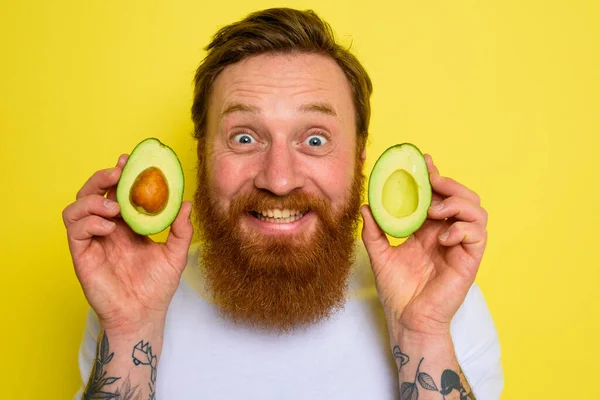 Sakallı ve dövmeli mutlu adam bir avokado tutuyor. — Stok fotoğraf