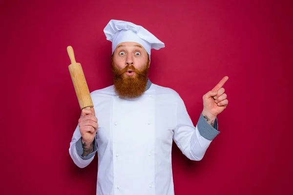 턱수염을 기르고 붉은 앞치마를 입은 놀란 요리사가 나무로 만든 전단을 들고 있다 — 스톡 사진