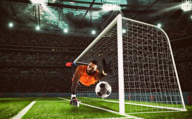 Turuncu üniformalı kaleci futbol maçı sırasında topu stadyumda yakalıyor.