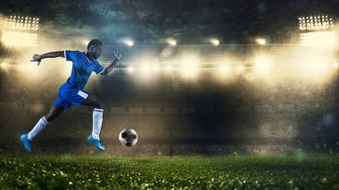 Mavi üniformalı futbolcu stadyumda topla hızla koşuyor.