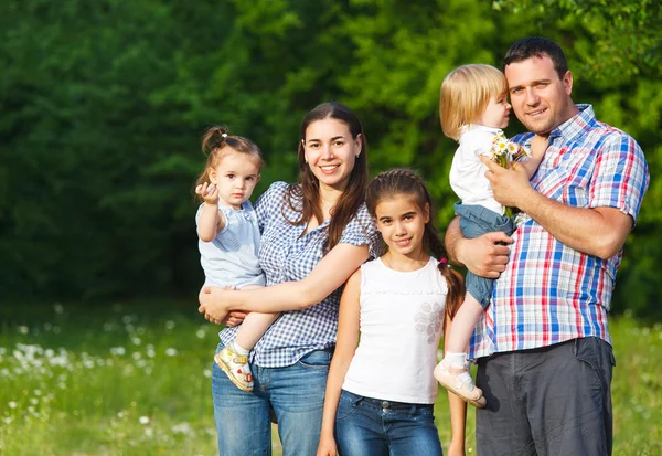 A family stands together in a rustic village setting with stone walls and houses. The scene is framed by vibrant purple flowers, adding a splash of color. The family consists of two adults and a child, all dressed in light, casual clothing, and they appear happy and relaxed.