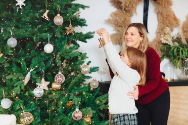 Neşeli bir anne ve güzel bir kız çocuğu rahat örgü kazaklarla Noel ağacını süslüyor yeni yıl mücevherleri ve oyuncaklarla dolu oturma odasında, gülümseyerek kış tatili atmosferini hissediyorlar. Aile tatilleri